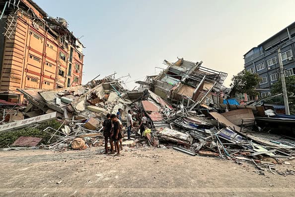 People stand past the debris of a collapsed building in Mandalay