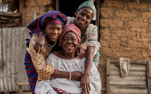 Three African women of different generations embracing and posing together outside a mud-brick building.