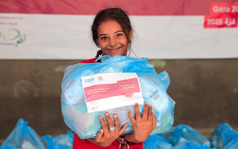 A smiling young girl in Gaza holds a large blue bag of emergency supplies for IDP families.