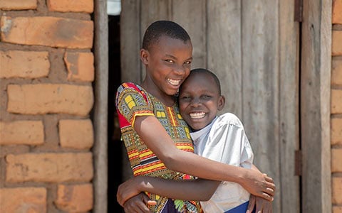 Two children smiling and hugging in front of a brick building and a wooden door.